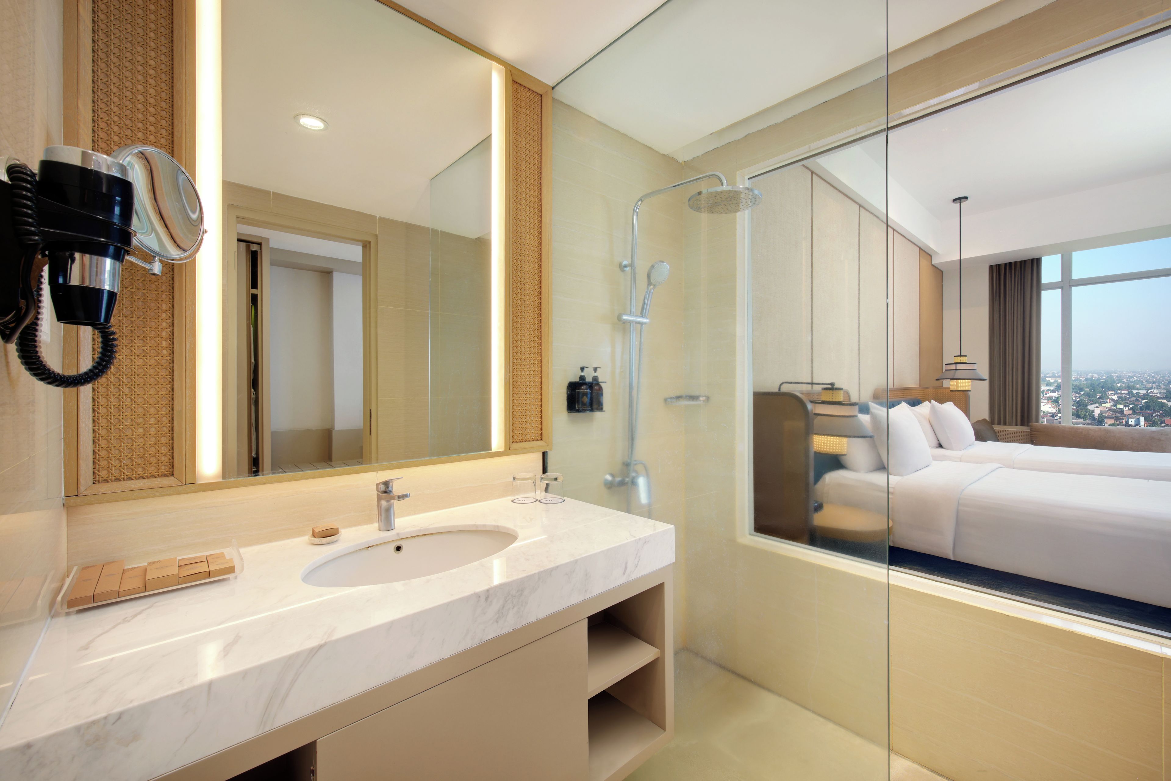 A modern hotel bathroom with a marble sink, a large mirror, a hairdryer, a shower, and a view of the city through a glass wall.