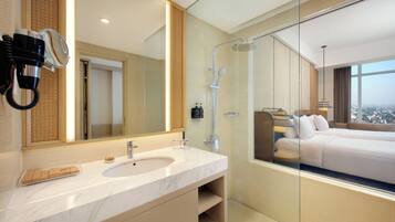 A modern hotel bathroom with a marble sink, a large mirror, a hairdryer, a shower, and a view of the city through a glass wall.