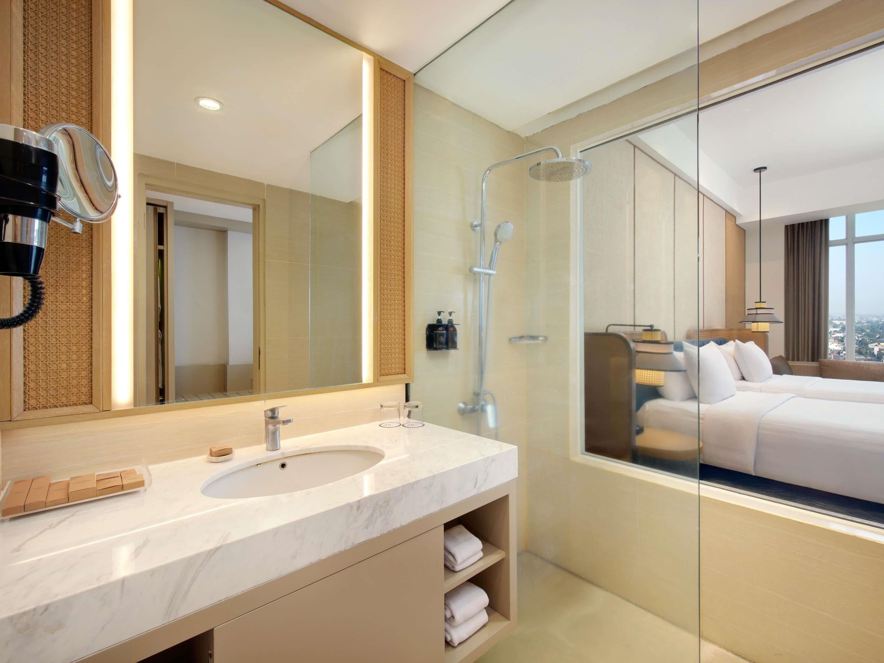 A modern bathroom with a large mirror, a shower, and a bed visible through the glass partition.