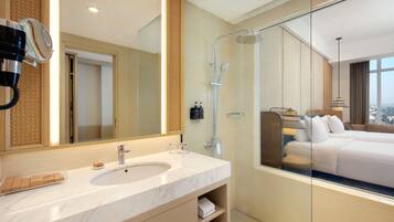 A modern bathroom with a large mirror, a shower, and a bed visible through the glass partition.