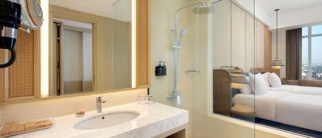 A modern bathroom with a large mirror, a shower, and a bed visible through the glass partition.