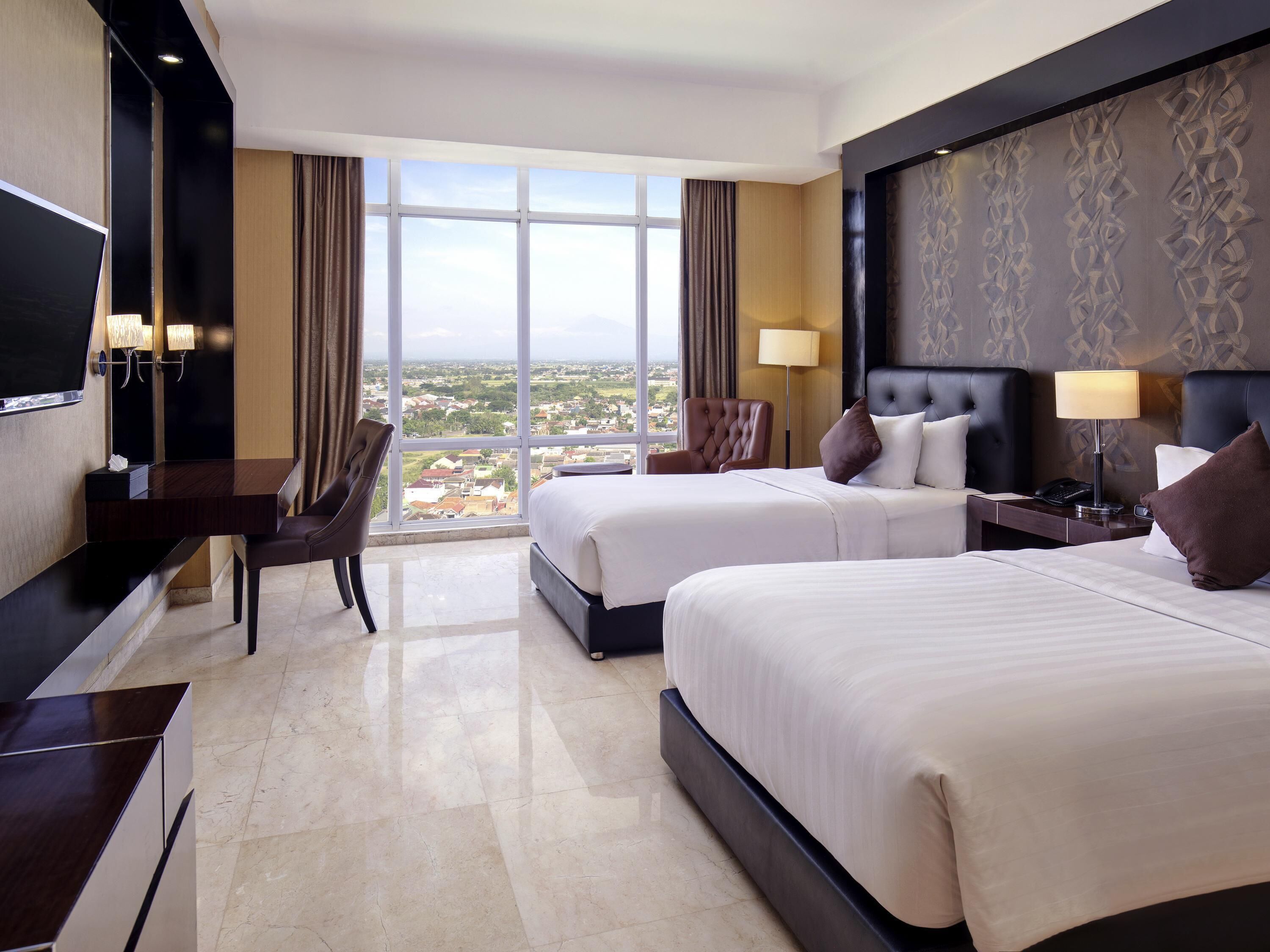 A hotel room with two beds, a desk, a chair, a TV, and a large window offering a city view.