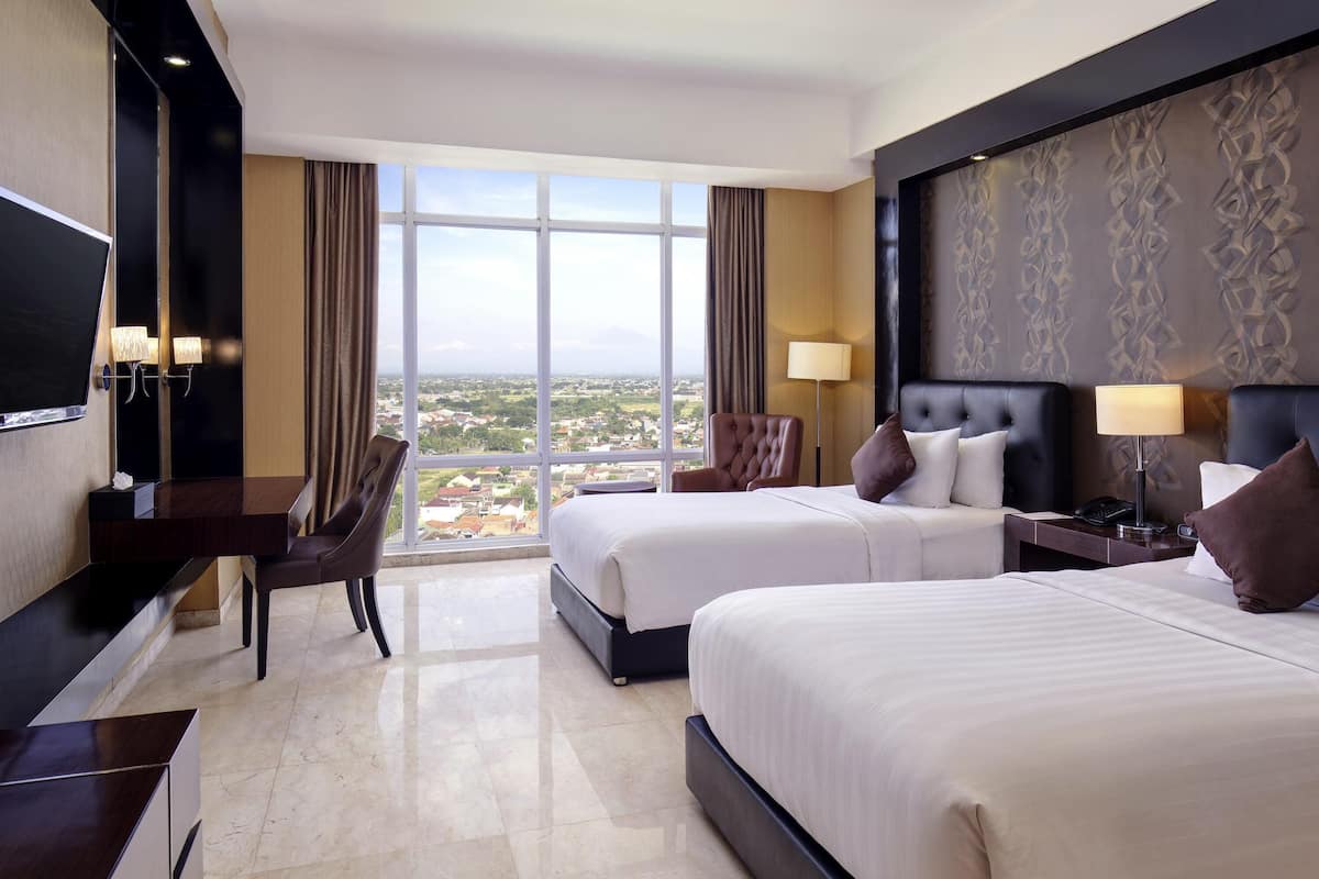A hotel room with two beds, a desk, a chair, a TV, and a large window offering a city view.