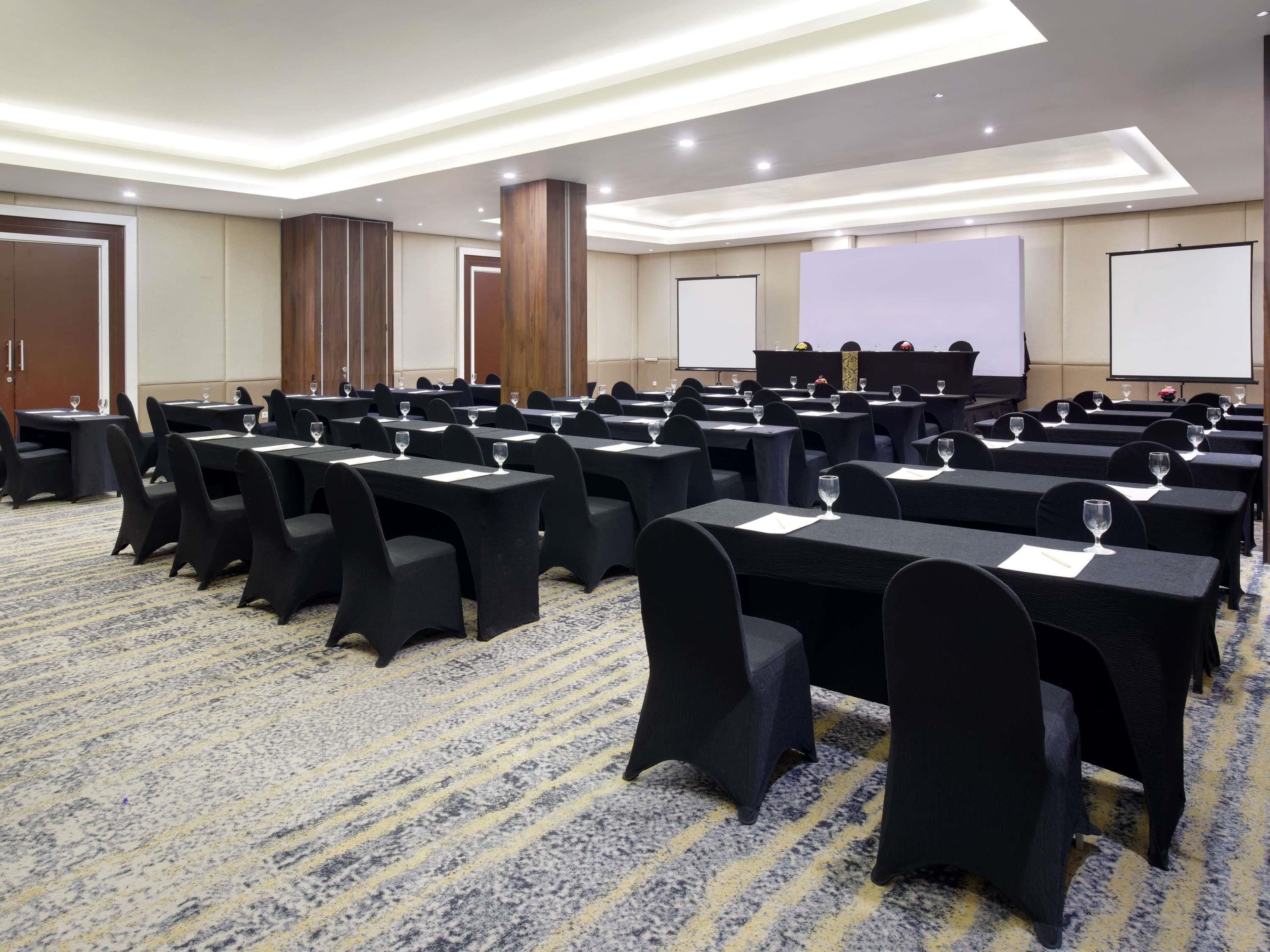Une salle de conférence avec des rangées de tables et de chaises, équipée de projecteurs et d’écrans.