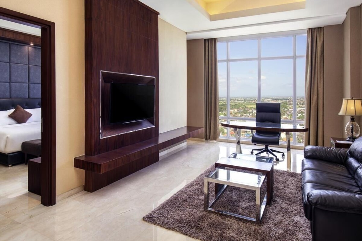 A modern hotel room with a flat-screen TV, a sofa, a dining table, and a large window offering a city view.