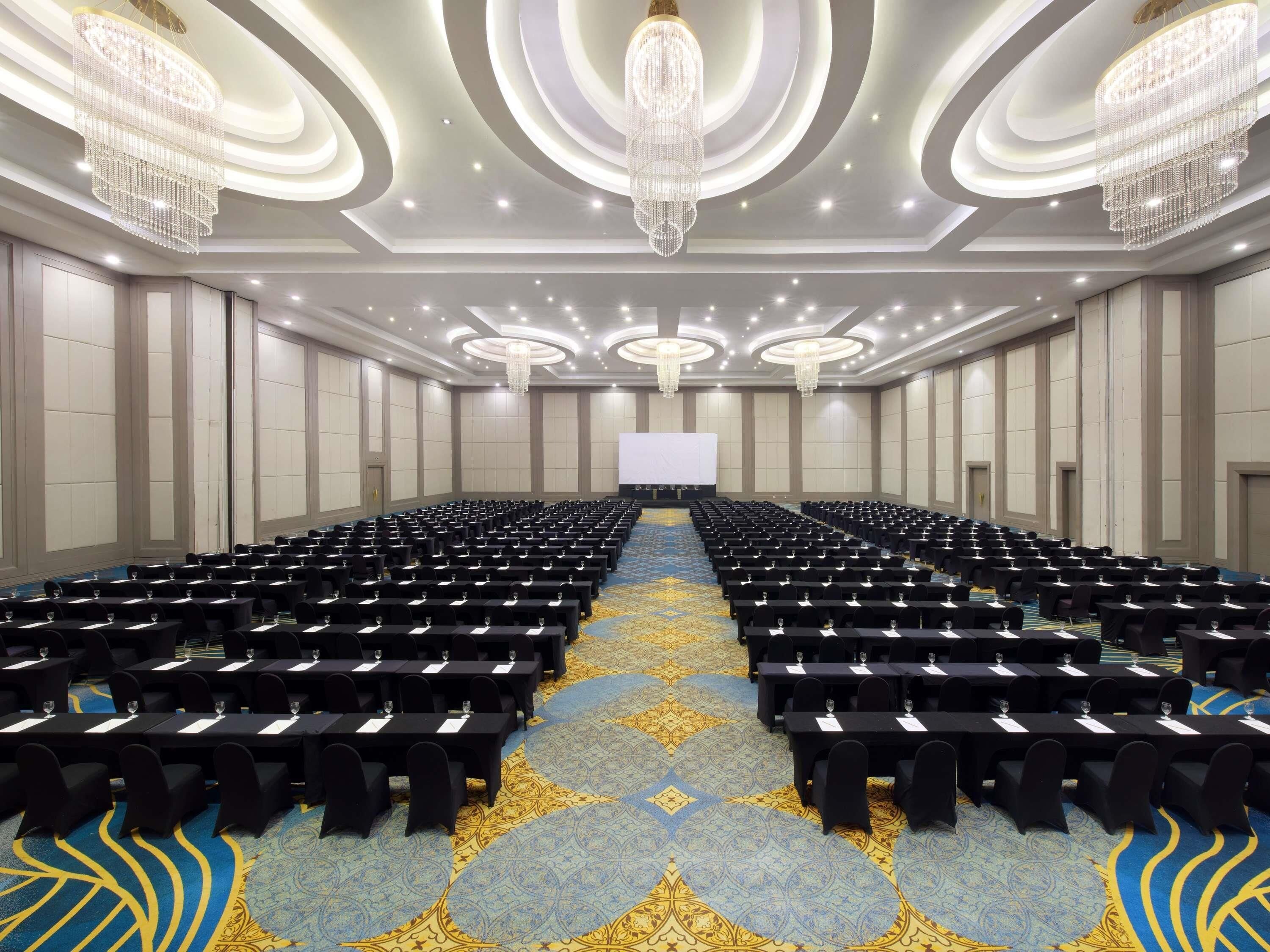 Un salón de conferencias con filas de mesas y sillas, una pantalla grande al frente y candelabros en el techo.