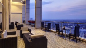 A modern rooftop lounge with wicker furniture, a glass railing, and a city view. - Grand Mercure Solo Baru (Grogol)