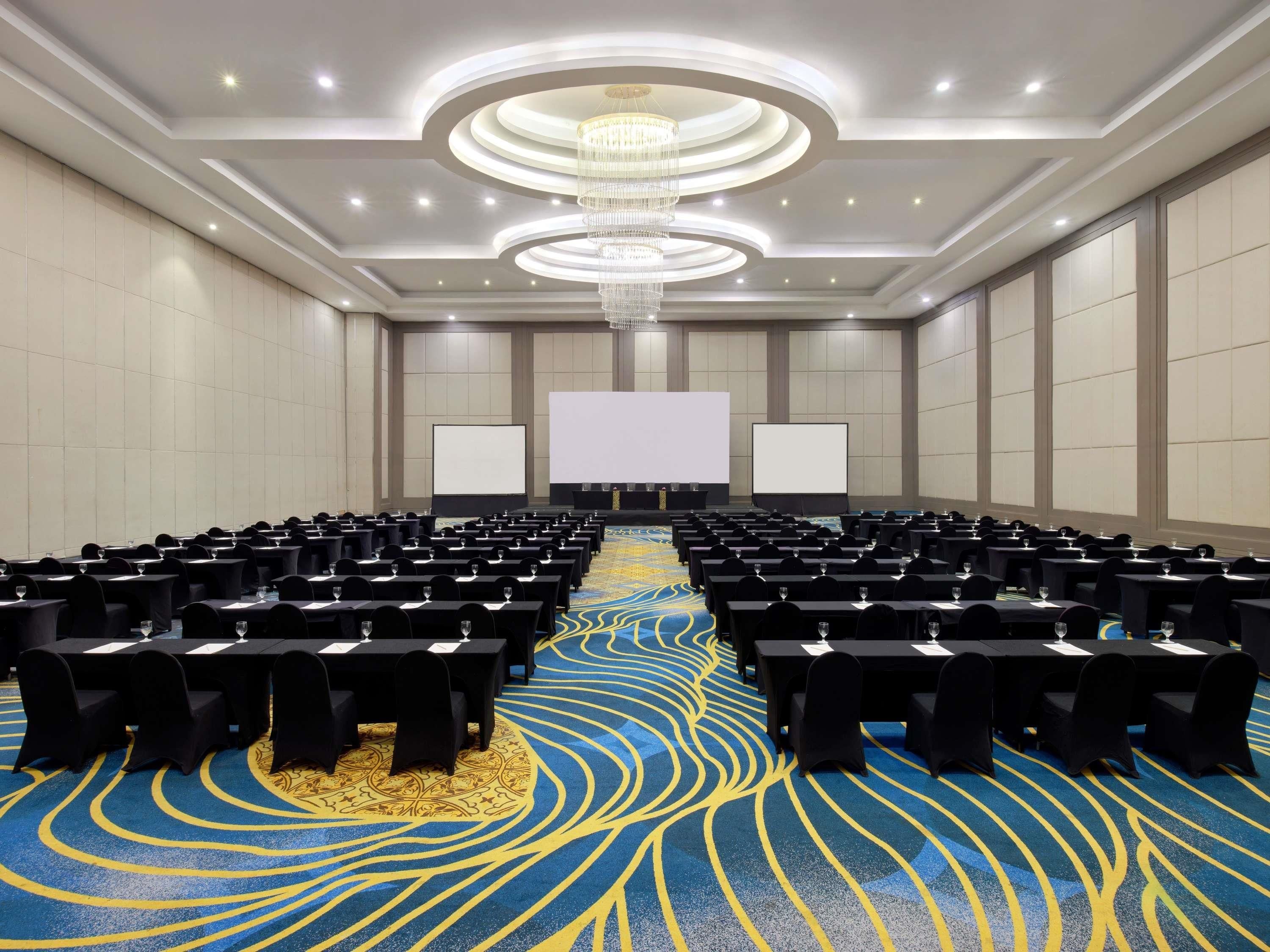 A conference room with rows of tables, a large screen, and a chandelier.