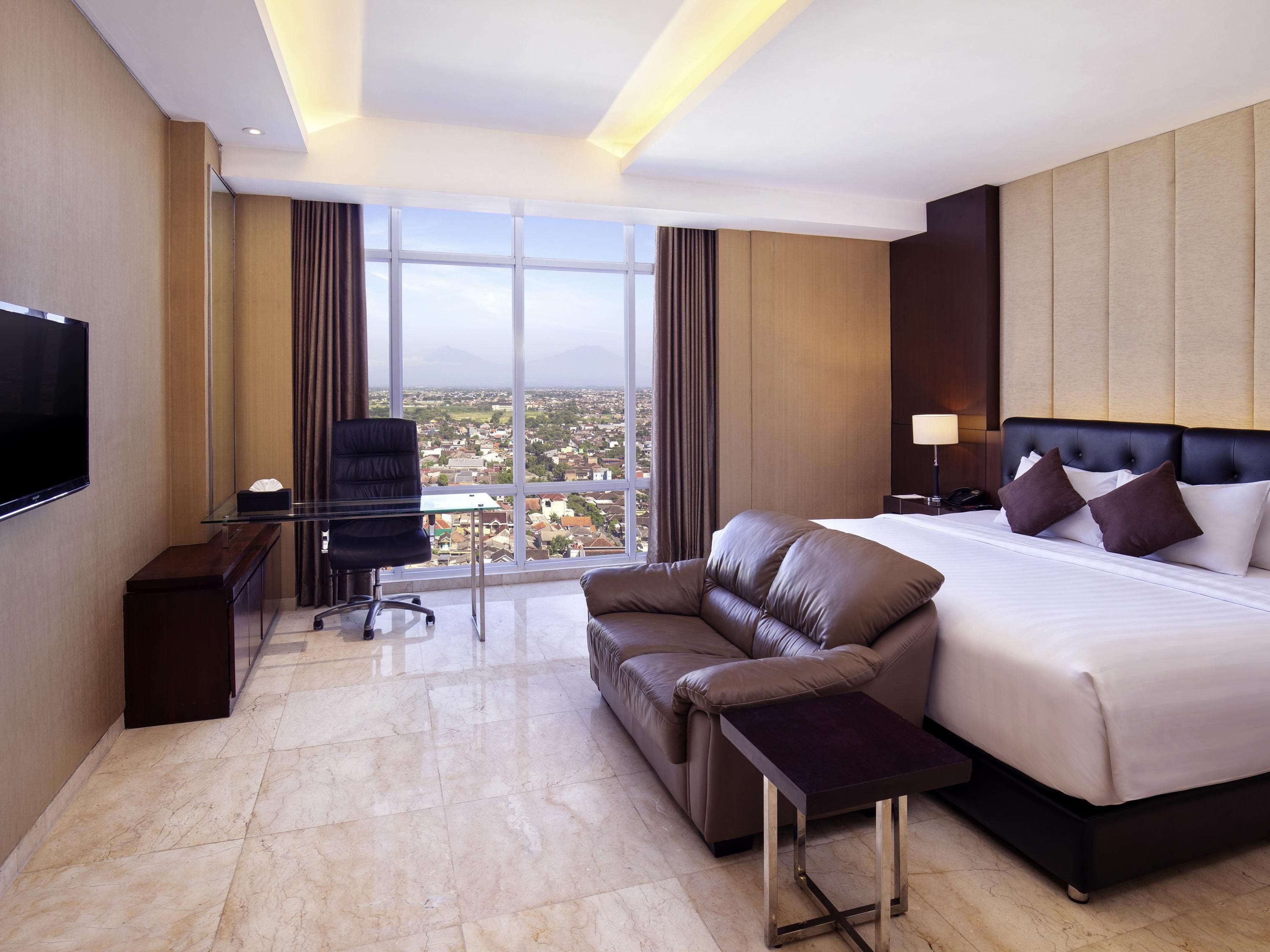 A hotel room with a large bed, a leather sofa, a desk with a chair, a TV, and a view of the city through large windows.