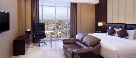 A hotel room with a large bed, a leather sofa, a desk with a chair, a TV, and a view of the city through large windows.