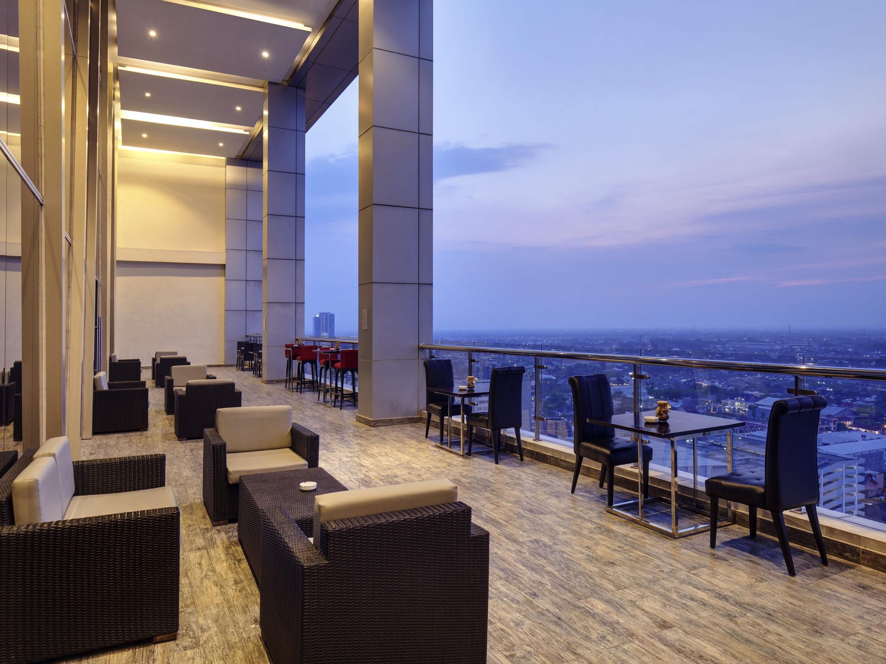 Un lounge panoramico moderno con mobili in vimini, ringhiera in vetro e vista sulla città.
