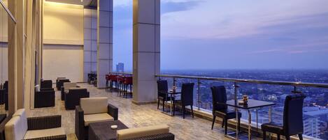 Un lounge panoramico moderno con mobili in vimini, ringhiera in vetro e vista sulla città.