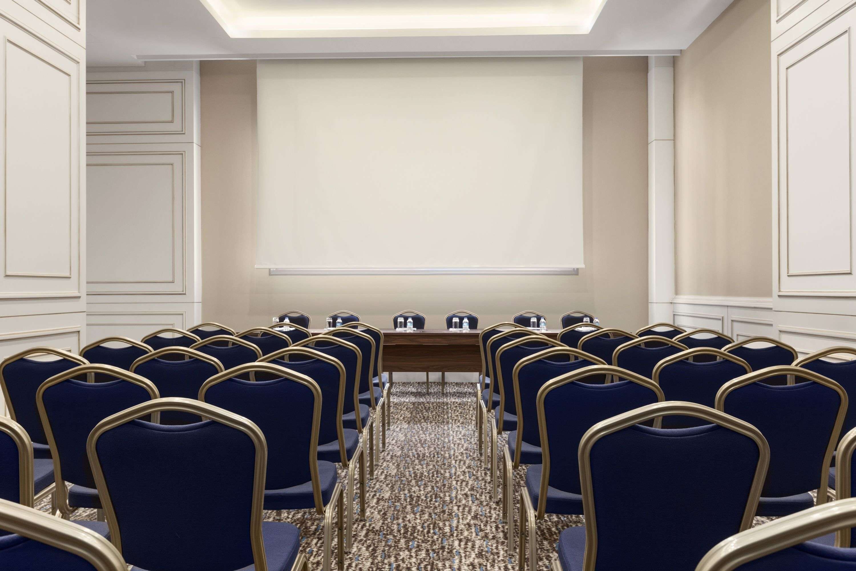 A conference room with rows of blue chairs, a long table with water bottles, and a projector screen.