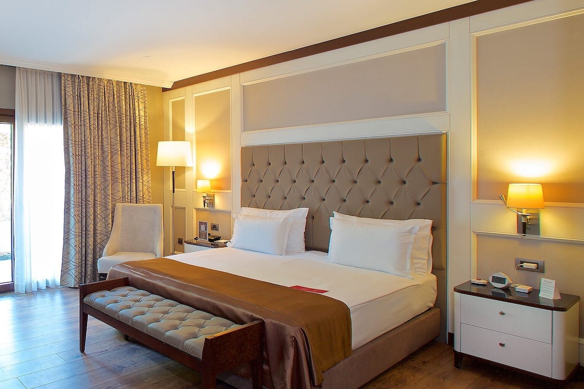A spacious hotel room with a large bed, a bench, two bedside tables with lamps, and a view of the outdoors through a large window.