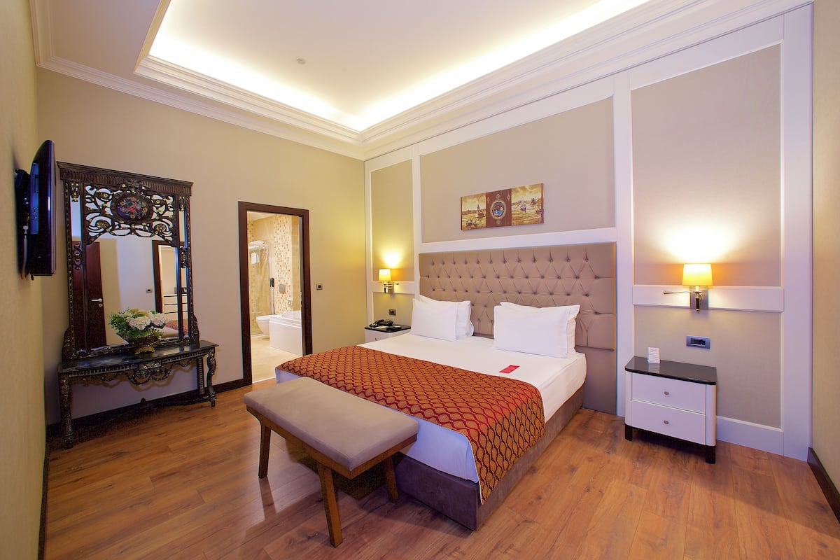 A hotel room with a large bed, bedside tables, a bench, a mirror, and a wall-mounted artwork.