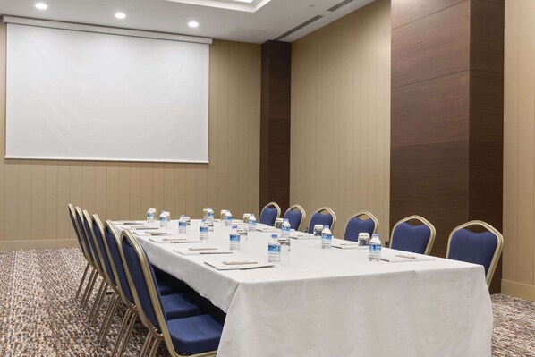 Un salón de conferencias con una larga mesa, sillas azules, botellas de agua y una pantalla para proyector.