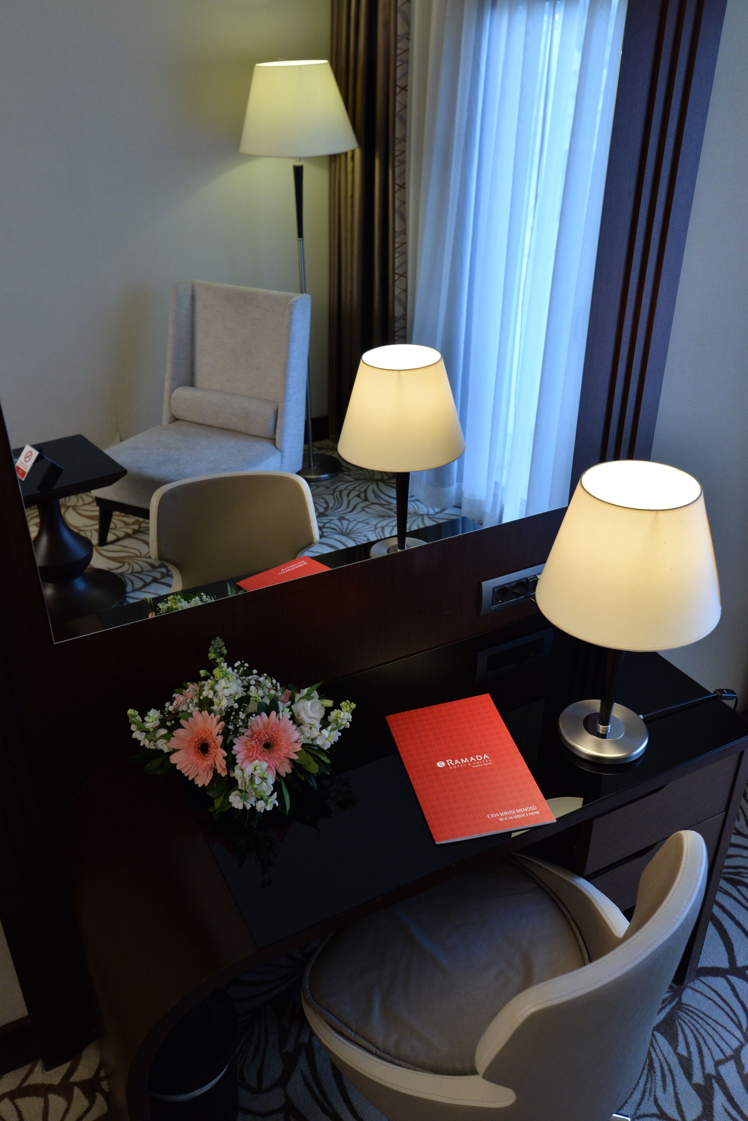 Una habitación de hotel con escritorio, dos lámparas, una silla, un espejo, un folleto rojo y un arreglo floral.