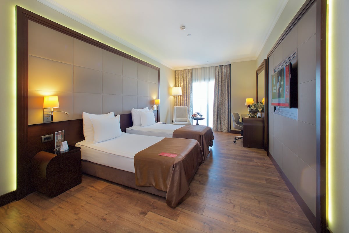 A hotel room with two beds, a desk, a chair, a TV, and a small table with a lamp.