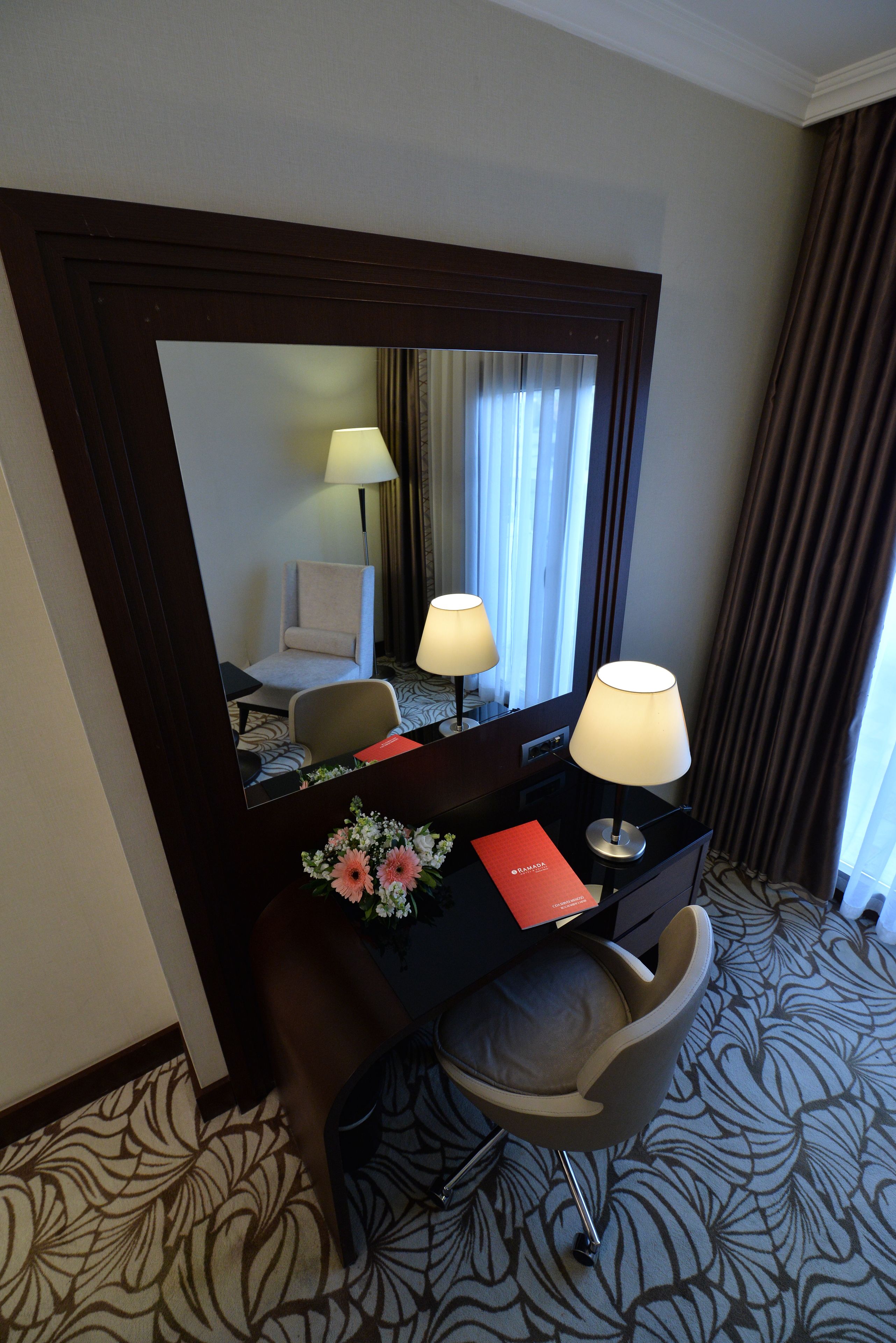 A hotel room with a desk, two lamps, a chair, a mirror, a red booklet, and a floral arrangement.