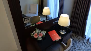 A hotel room with a desk, two lamps, a chair, a mirror, a red booklet, and a floral arrangement.