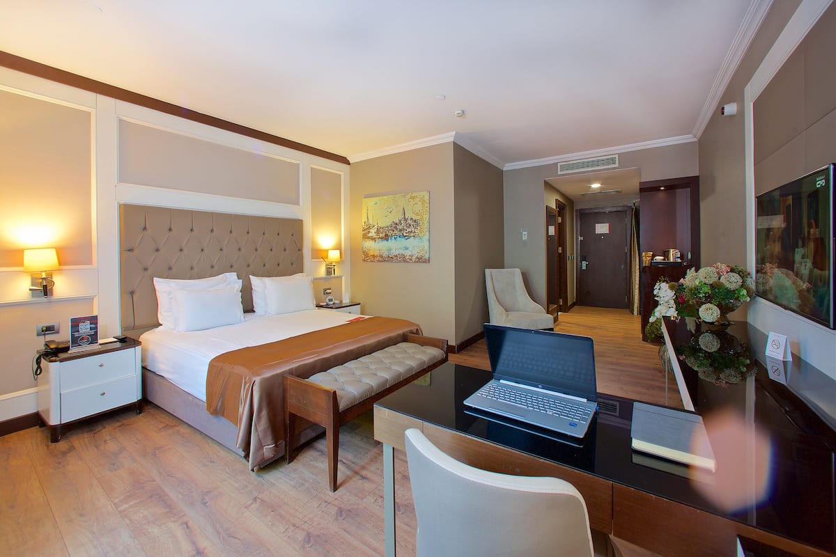 A hotel room with a bed, a desk with a laptop, a chair, a nightstand, and a lamp.