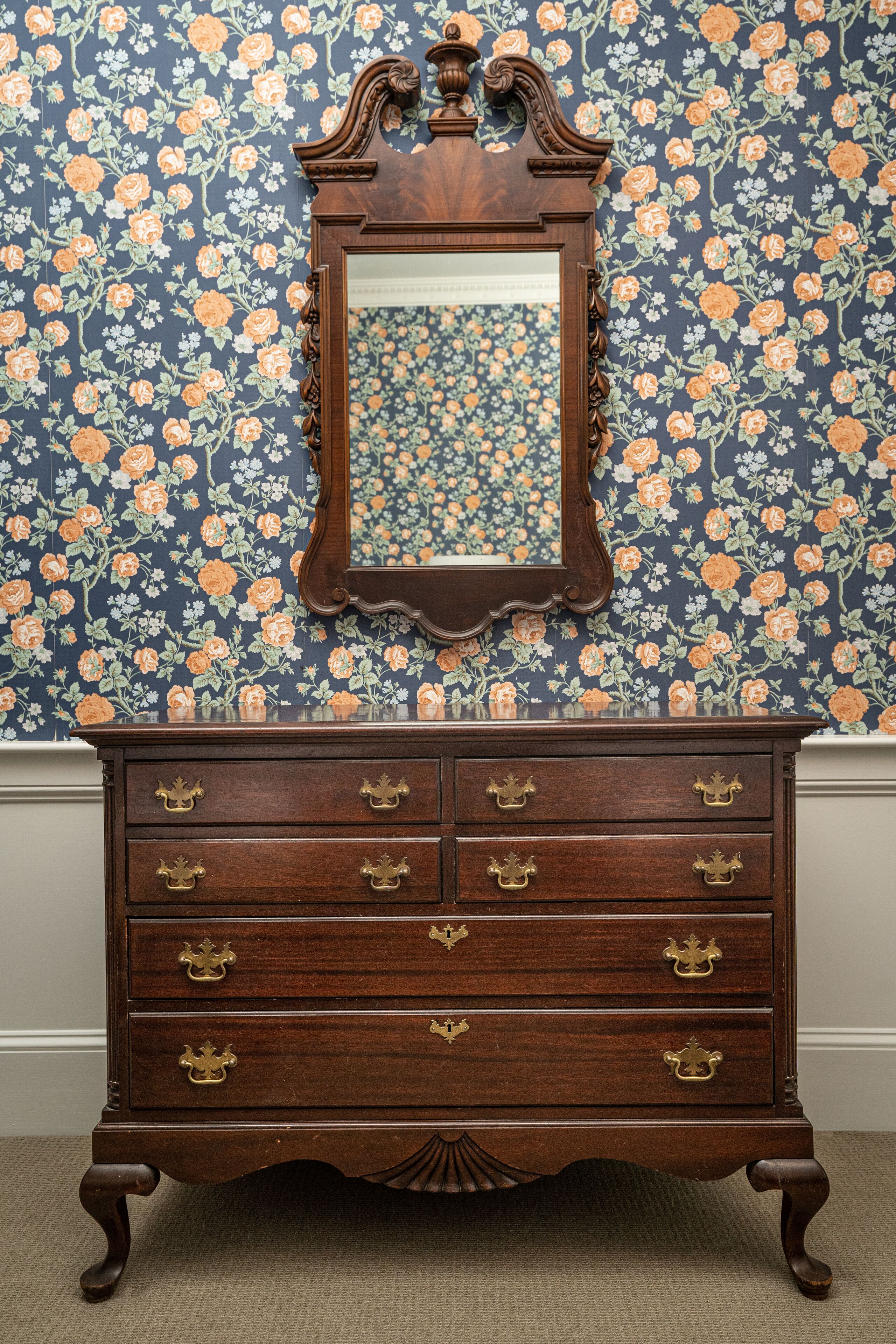 A wooden dresser with ornate carvings and brass handles against a floral wallpaper background.
