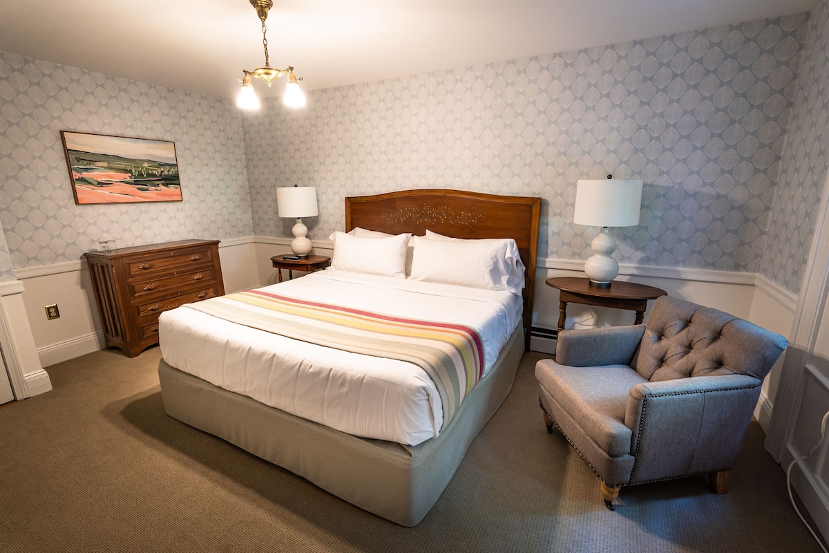 A neatly arranged bedroom with a large bed, two bedside tables with lamps, a wooden dresser, and a tufted armchair.