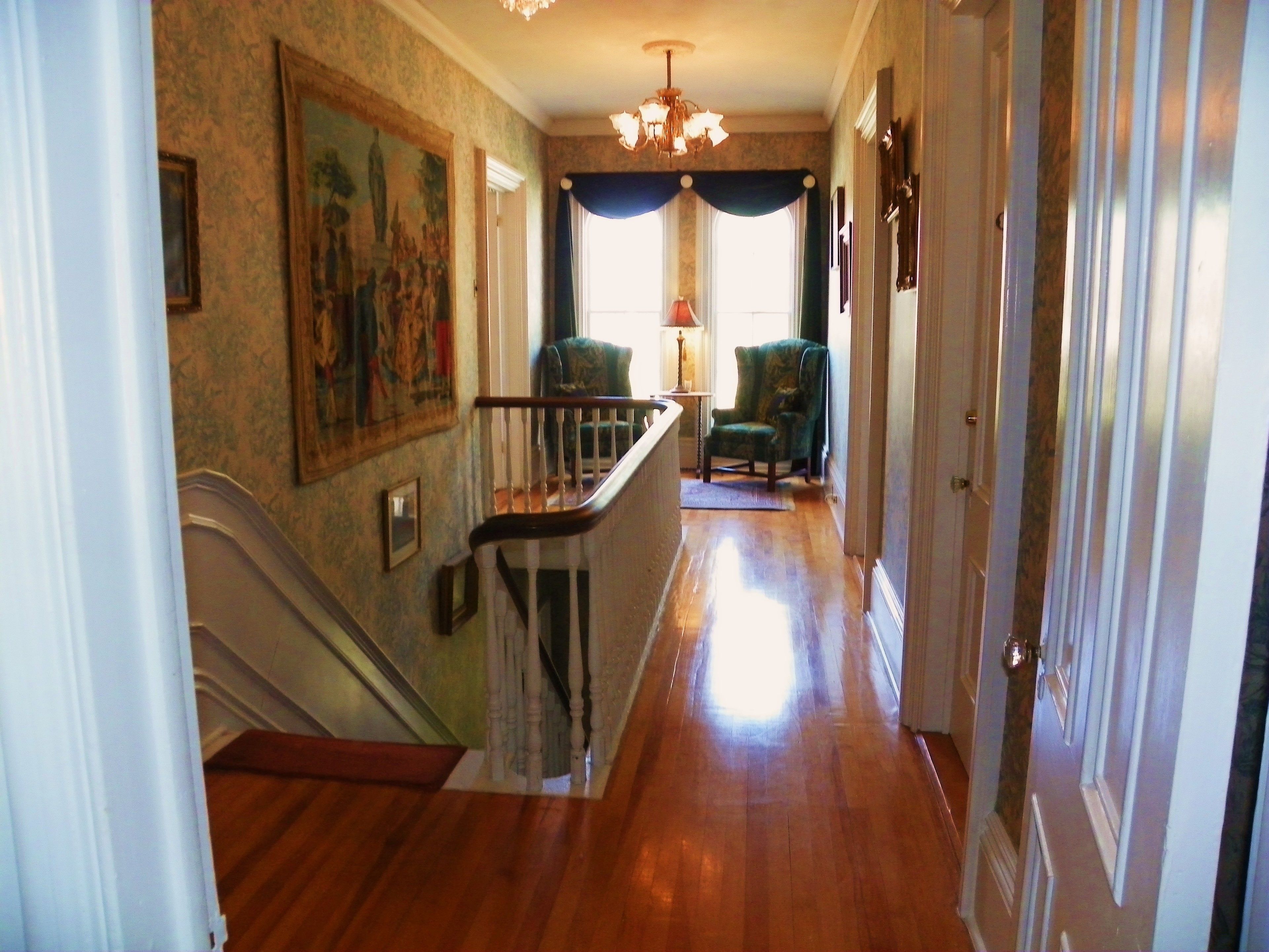 A hallway with a wooden staircase, a chandelier, a painting on the wall, and a chair.