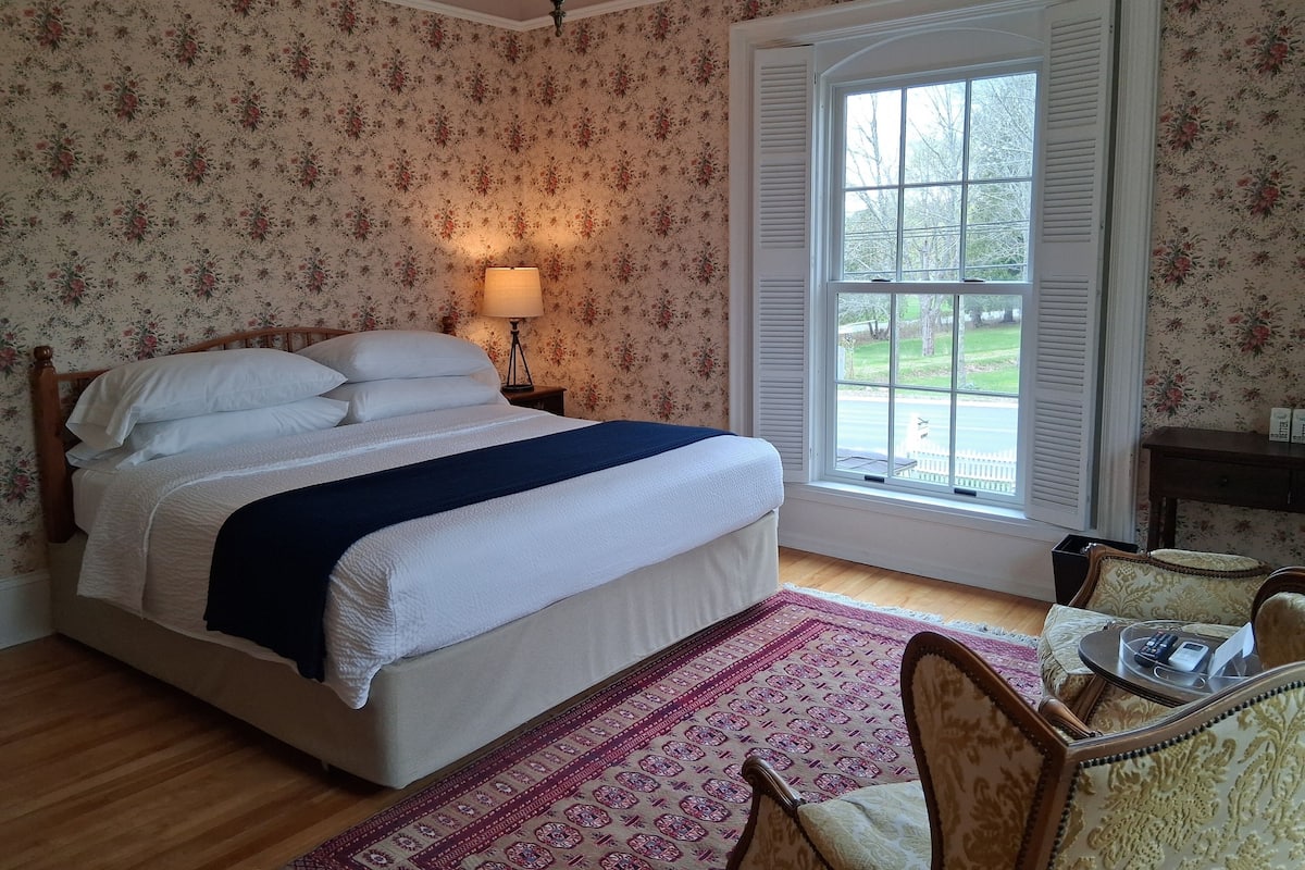 A neatly arranged bedroom with a bed, bedside lamp, and a view of the outdoors through a window.