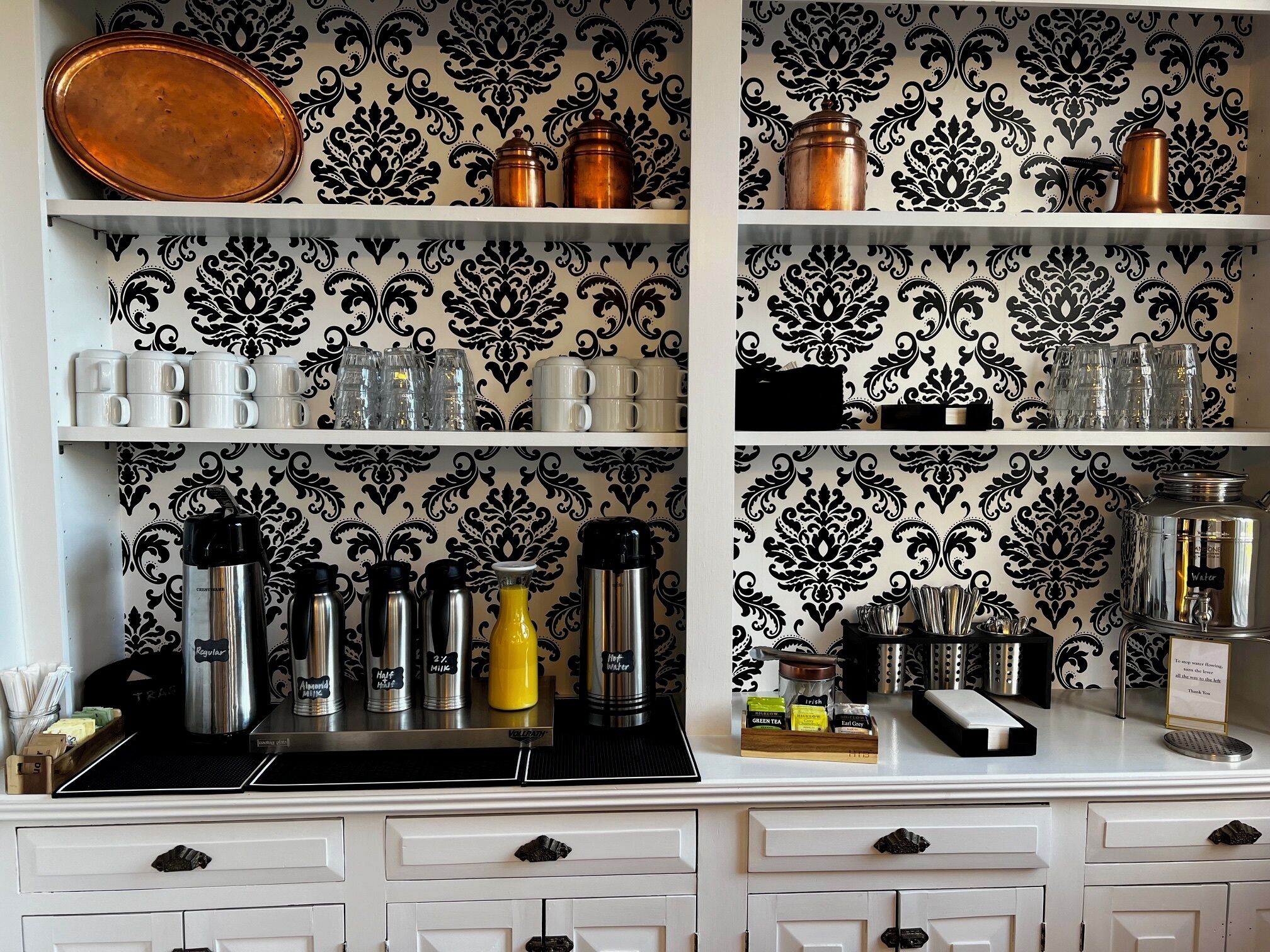 A kitchen with white cabinets, black and white patterned wallpaper, and various kitchen items like mugs, thermoses, and a coffee machine.