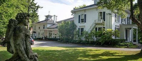 A stately white building with a prominent entrance and a balcony, surrounded by lush greenery.