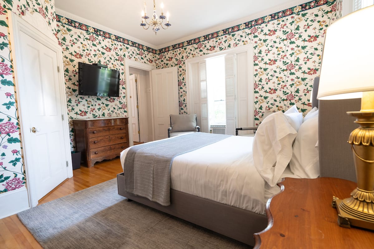 A bedroom with floral wallpaper, a bed, a dresser, a TV, and a lamp.