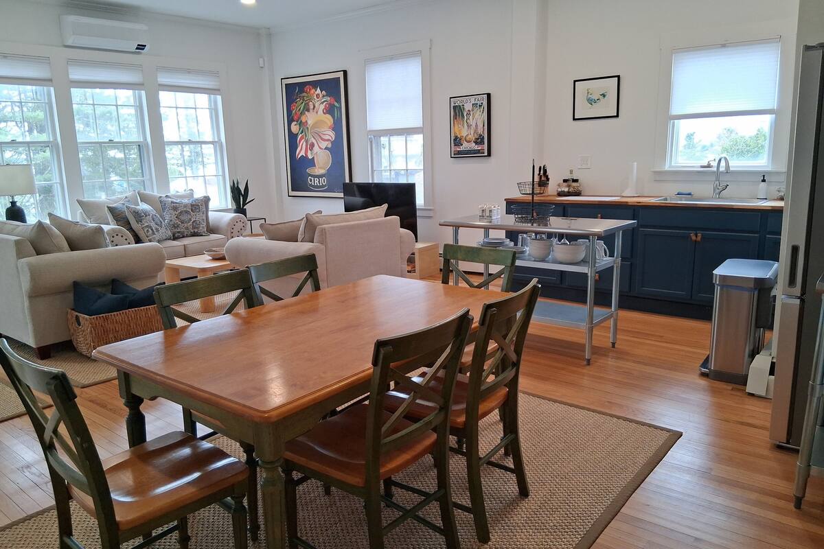 A spacious living room with a wooden dining table and chairs, a sofa set, and a kitchen area with a sink and cabinets.