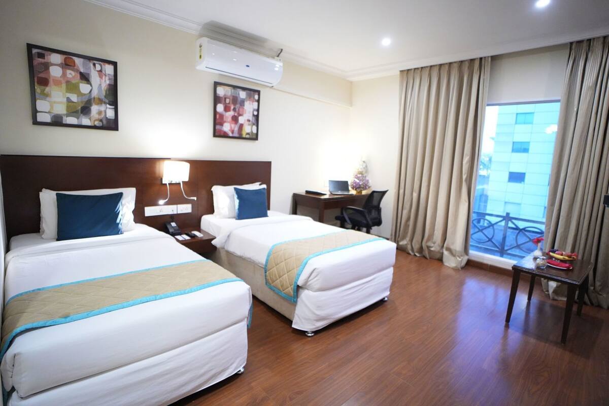 A hotel room with two beds, a desk, a chair, an air conditioner, and a window with curtains.