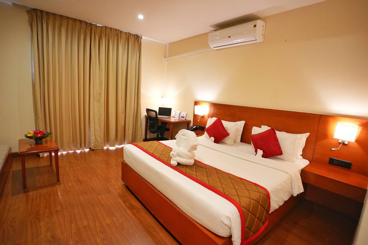 A hotel room with a bed, a desk, a chair, a lamp, and an air conditioning unit.