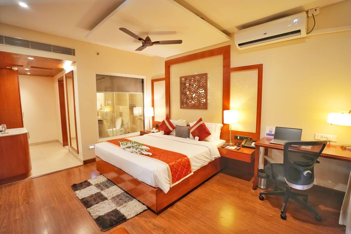 A hotel room with a large bed, a desk with a computer, a chair, a ceiling fan, and an air conditioning unit.