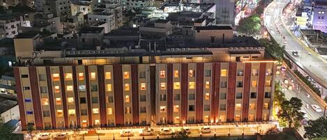 A multi-story hotel with a row of rooms, a parking area, and a busy street in the background.