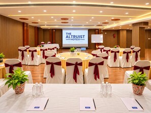 A conference room with round tables, white chairs with red sashes, a large screen displaying 'THE ALTRUIT HOTELS', and potted plants. - The Altruist Hitech Hotel (Hyderabad)