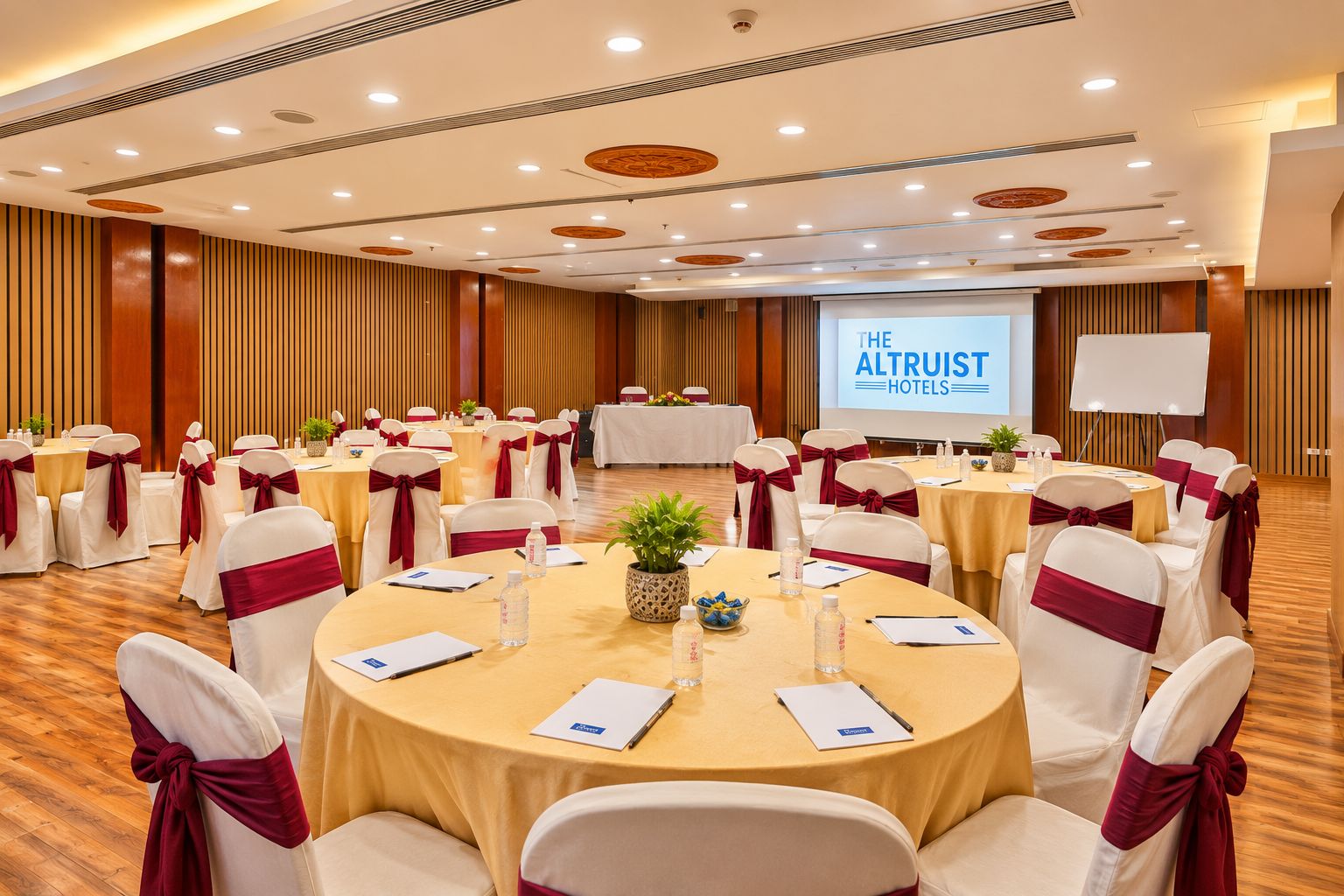 A conference room with round tables, chairs with red sashes, and a projection screen displaying 'THE ALTRUIT HOTELS'.