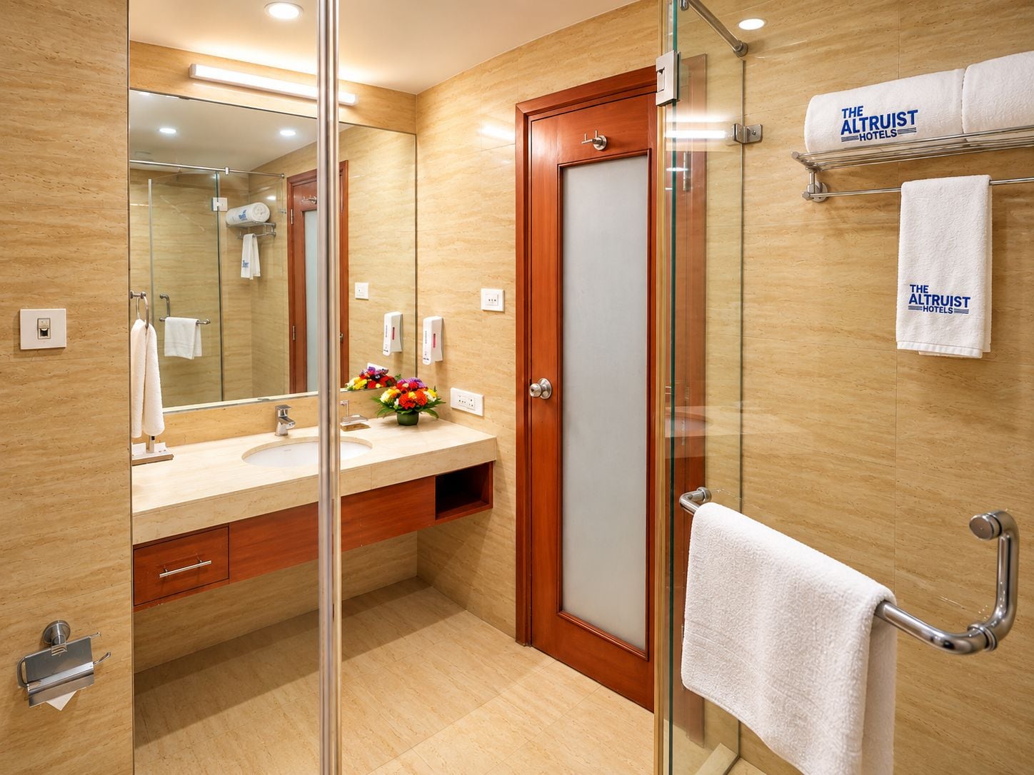 A hotel bathroom with a glass shower enclosure, a wooden vanity, a mirror, and a towel rack with branded towels.