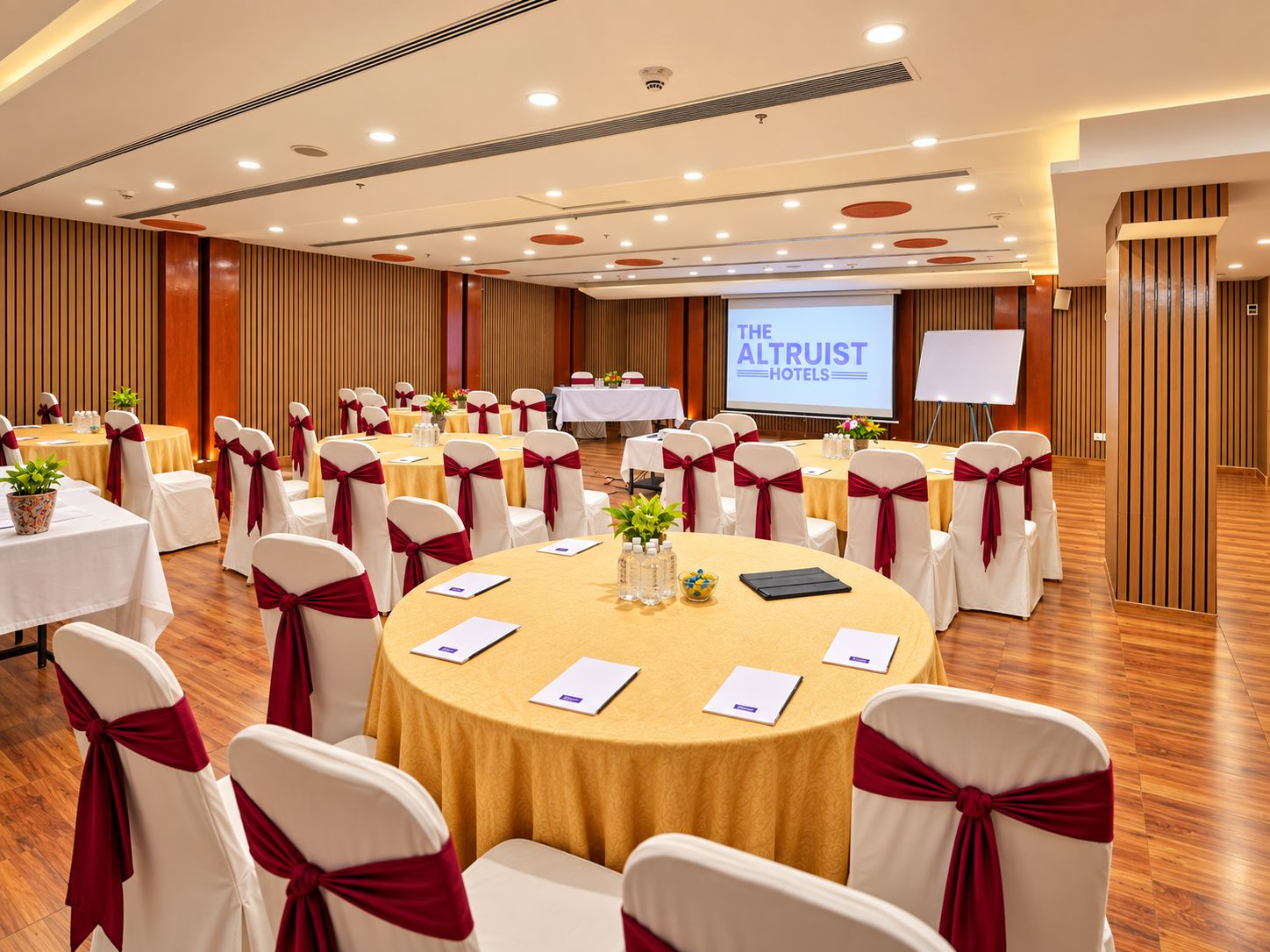 A conference room with round tables, white chairs with red sashes, and a projection screen displaying 'THE ALTRUIST HOTELS'.