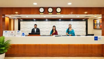 A reception desk with three staff members, clocks displaying different time zones, and signs indicating services and safety measures.
