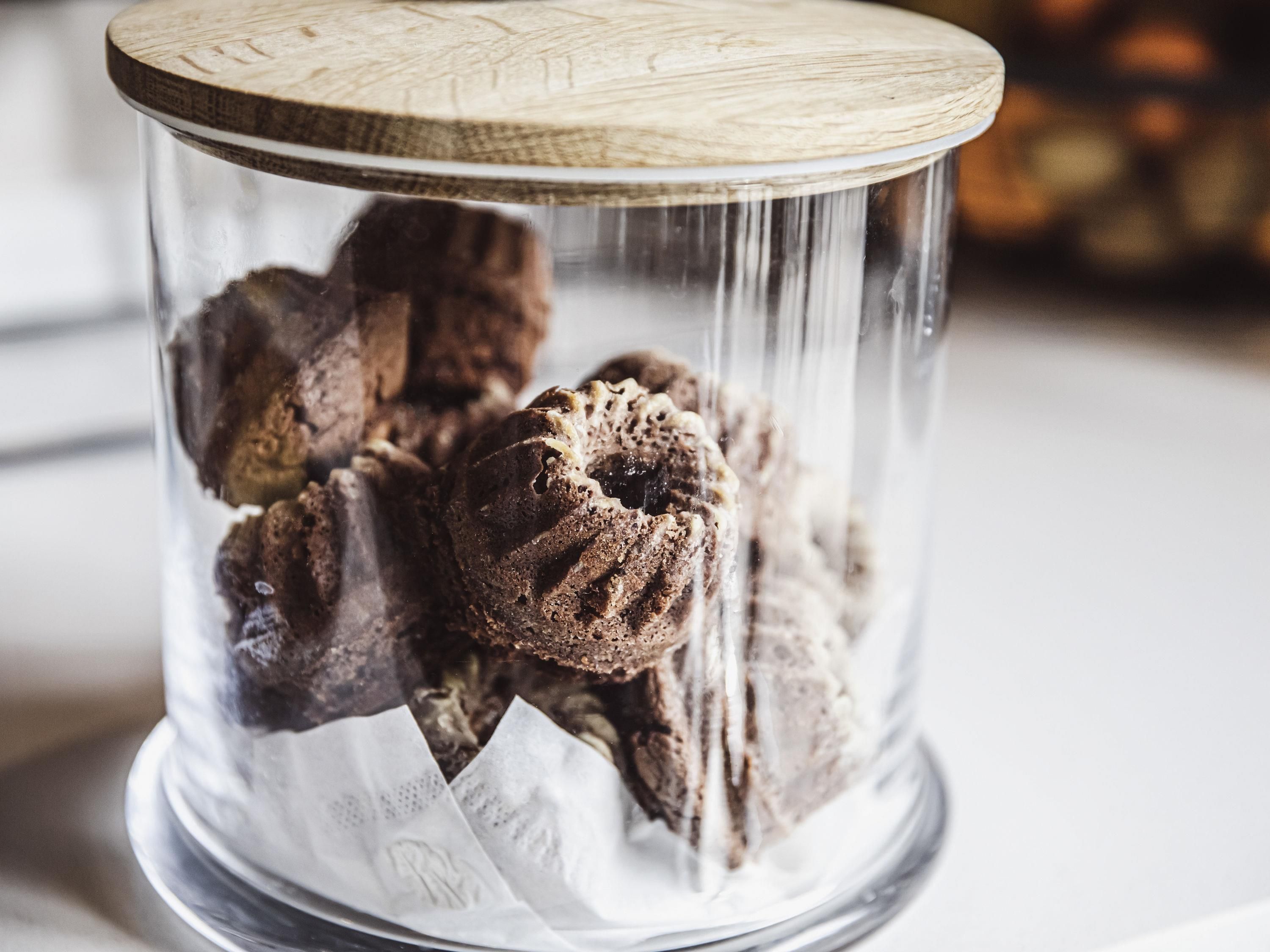 A glass container with a wooden lid holding chocolate cookies.