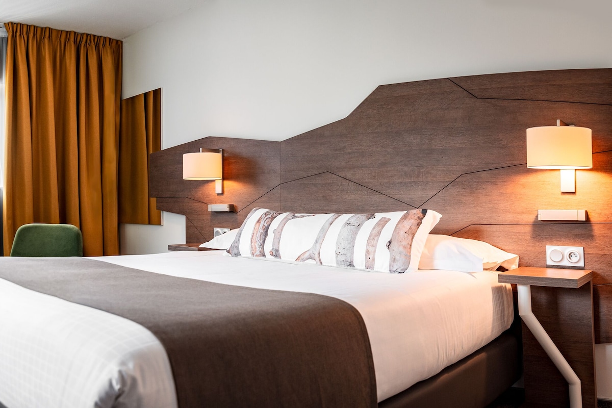 A hotel room with a large bed, a desk, a chair, two bedside lamps, and a window with curtains.