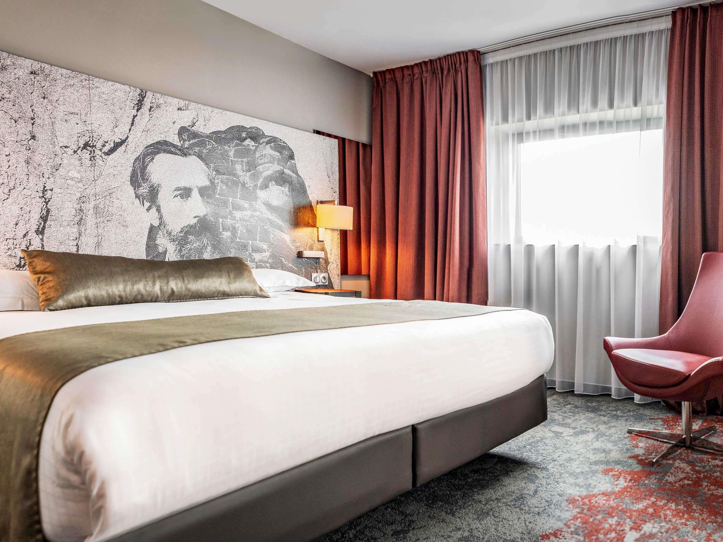 A hotel room with a large bed, a red chair, a wall-mounted lamp, and a window with curtains.