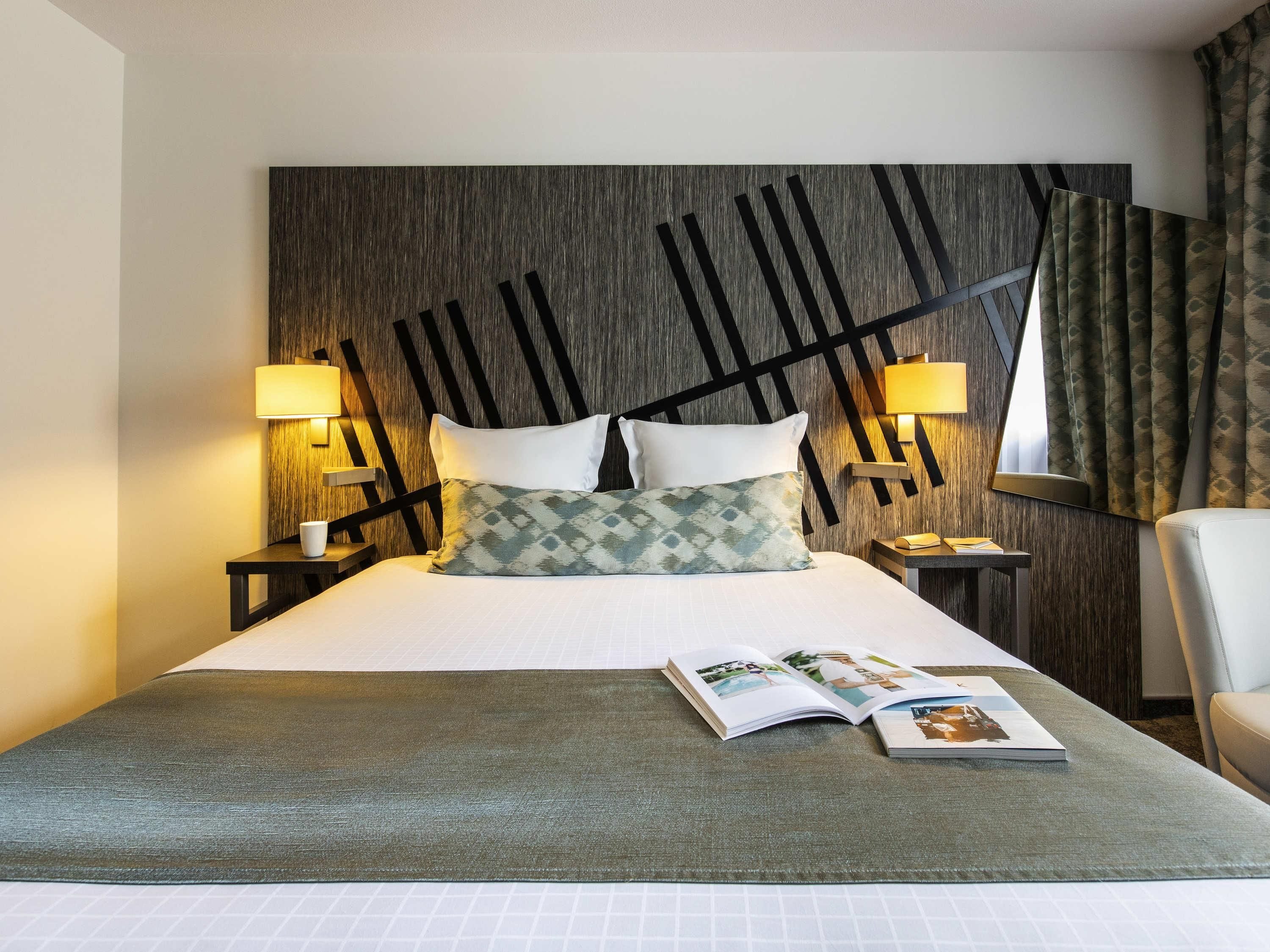 A hotel room with a bed, two bedside lamps, a chair, a window with curtains, and a magazine on the bed.