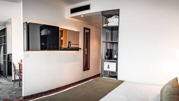 Una habitación de hotel moderna con sofá, una silla roja, una mesita, cama, armario y baño con ducha e inodoro.
