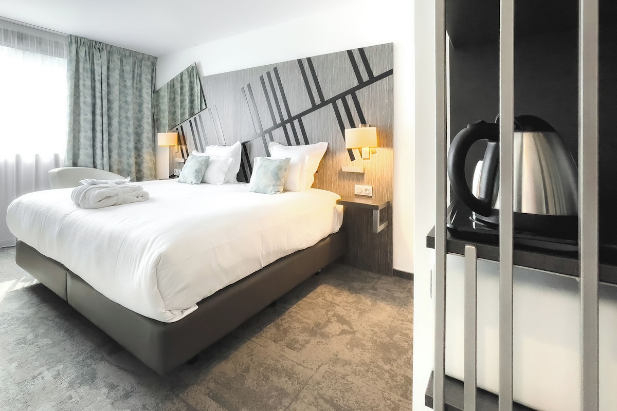 A modern hotel room with a large bed, a wardrobe, and a nightstand with a lamp.