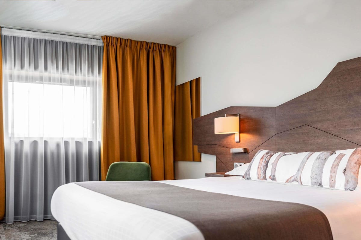 Ein Hotelzimmer mit einem großen Bett, einem grünen Sessel, einem Nachttisch mit Lampe und einem Vorhangfenster.