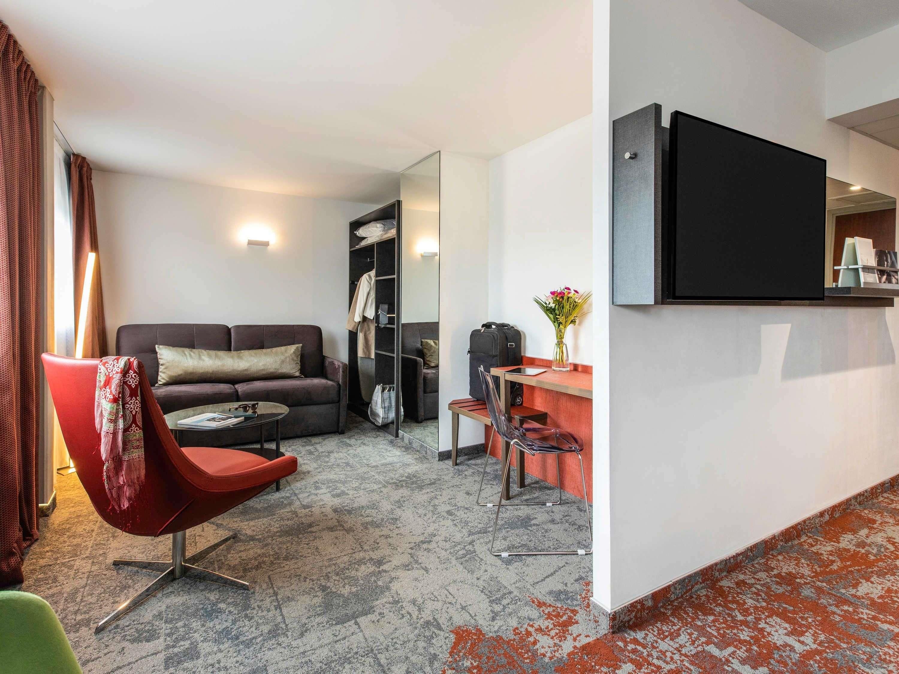 A modern hotel room with a sofa, a red chair, a small table, a desk, a mirror, and a TV.
